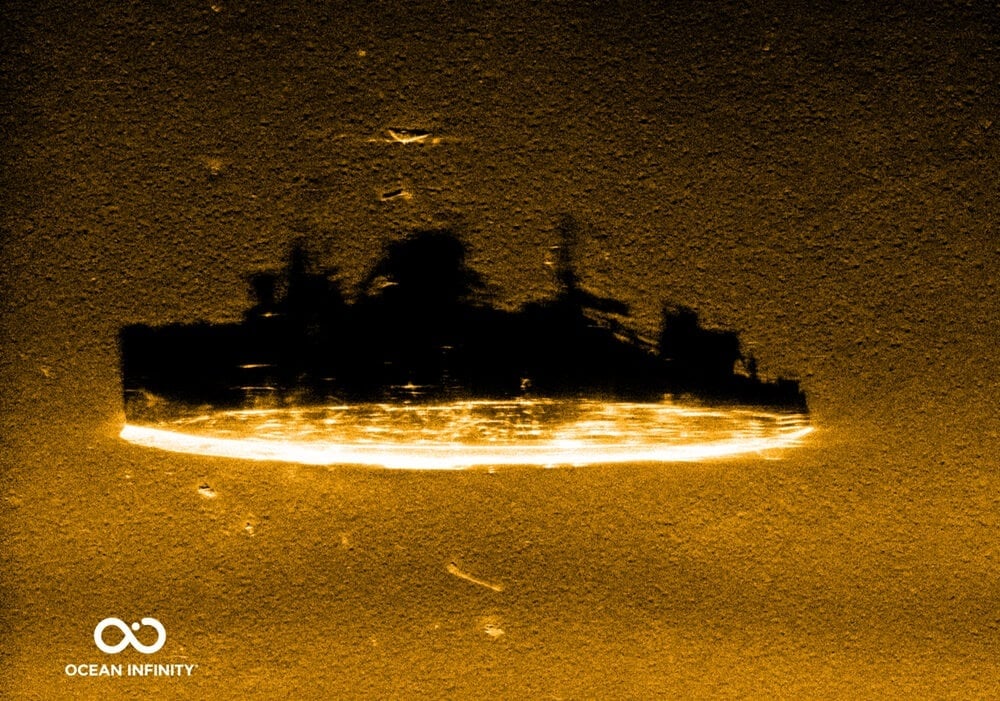 Sonar scan image of a sunken ship on the ocean floor. The ship appears as a dark silhouette against a golden background, with details of its structure faintly visible. The Ocean Infinity logo is in the bottom left corner.