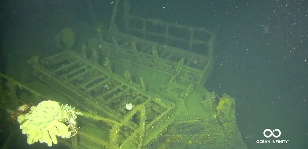 A submerged, algae-covered structure rests on the ocean floor. It's encrusted with marine life, hinting at a lengthy submersion. The image has a dark, murky ambiance, with visible sediment floating in the water. Ocean Infinity's logo is in the corner.