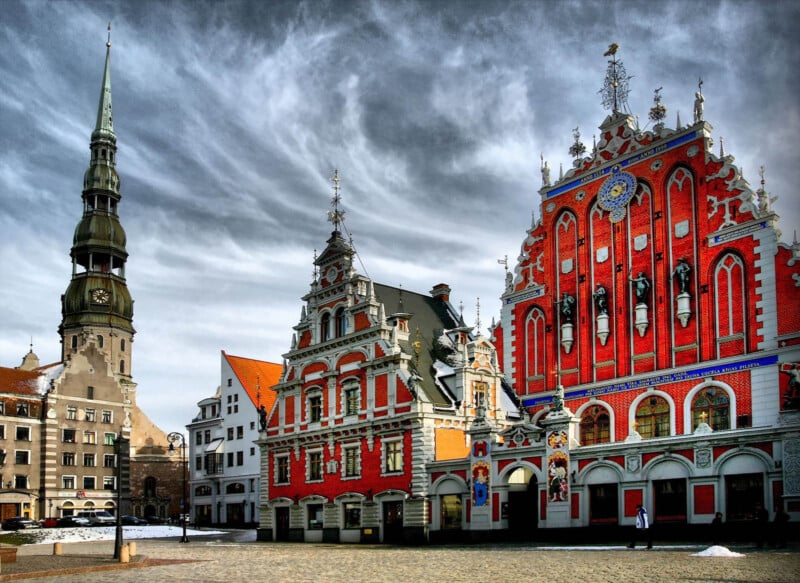 A historic European town square featuring ornate red brick buildings with decorative facades and a tall church spire. The sky is overcast, adding a dramatic backdrop to the intricate architecture.