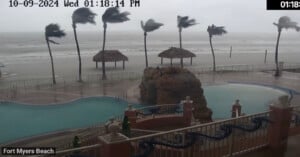 View of a stormy beach with strong winds bending palm trees. Waves crash near a rock formation by a pool. Cloudy sky and rain create a hazy atmosphere. Time and date displayed in the corner: October 9, 2024, 01:18 PM. Location: Fort Myers Beach.