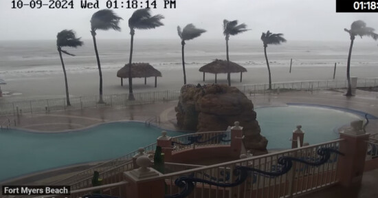 View of a stormy beach with strong winds bending palm trees. Waves crash near a rock formation by a pool. Cloudy sky and rain create a hazy atmosphere. Time and date displayed in the corner: October 9, 2024, 01:18 PM. Location: Fort Myers Beach.