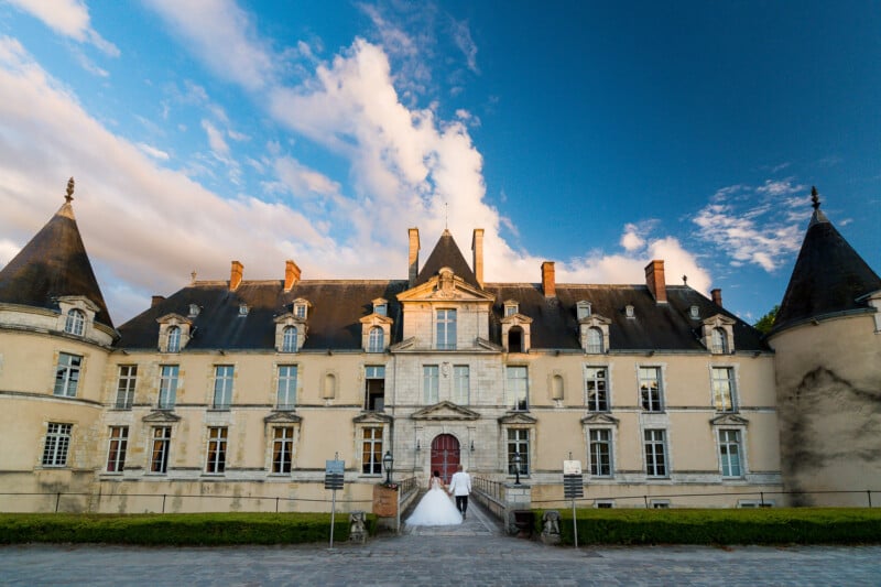 A couple walking hand in hand toward a grand, historic chateau with tall turrets and numerous windows. The sky is partly cloudy, casting a warm, golden light on the building's facade.