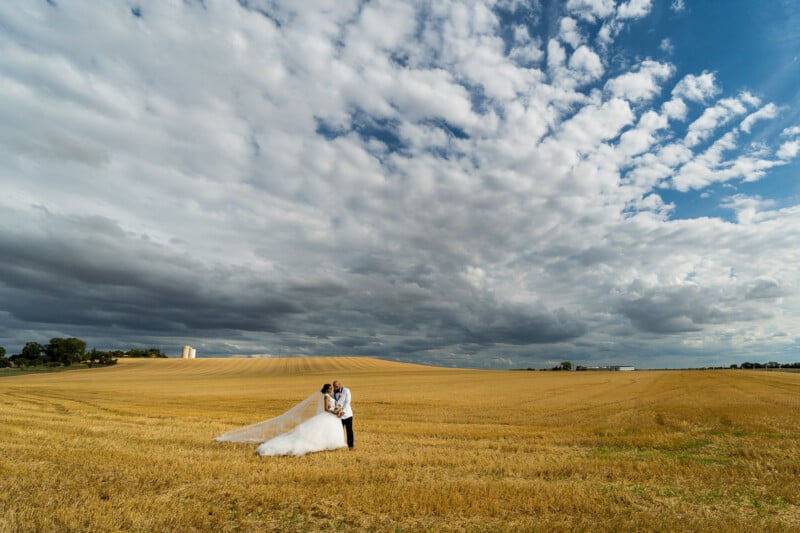 A bride and groom embrace in a vast, golden wheat field under a dramatically cloudy sky. The bride's long veil flows on the ground, and the horizon stretches with rolling fields and distant trees.