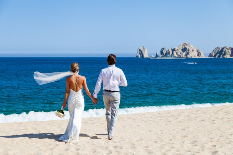 A bride and groom walk hand in hand on a sandy beach, facing the ocean. The bride wears a white gown with a veil blowing in the breeze, and the groom is in a white shirt and light pants. Rocky formations are visible in the distance under a clear blue sky.