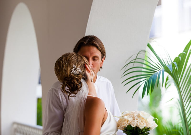 A couple shares a kiss in a white arched hallway. The woman is wearing a bridal gown with a veil and holding a bouquet of white roses. A palm frond is visible in the background.