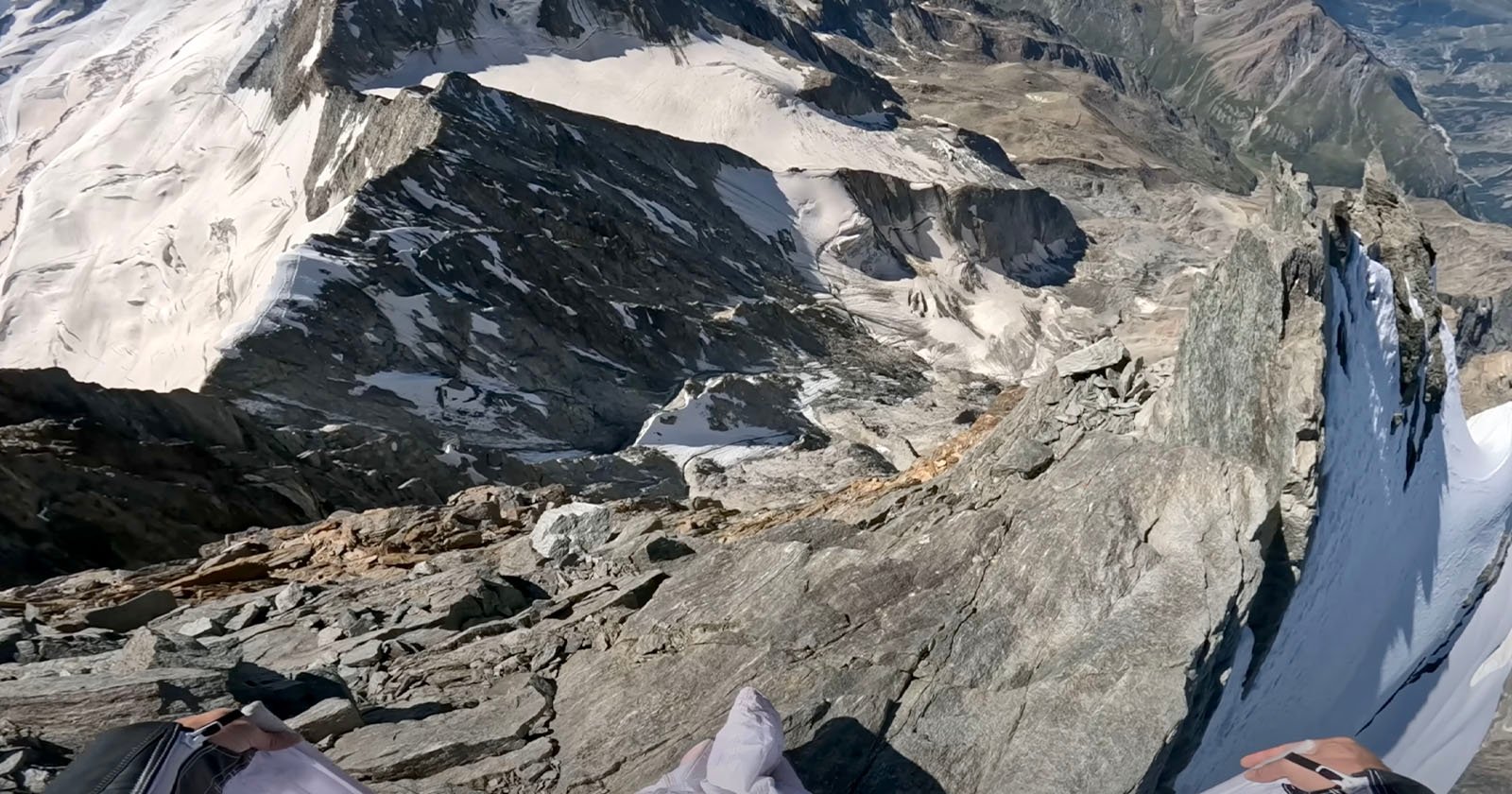 A breathtaking view from a high mountain peak, showing jagged rocky slopes and patches of snow. The horizon displays towering peaks and a distant valley under a clear blue sky. A climber’s feet are visible, resting on the rocks.