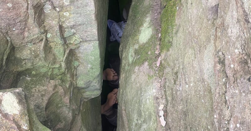 A narrow crevice between large mossy rocks reveals a glimpse of three feet, suggesting people are squeezing through or resting within the rock formation. Scattered shoes are visible in the background.