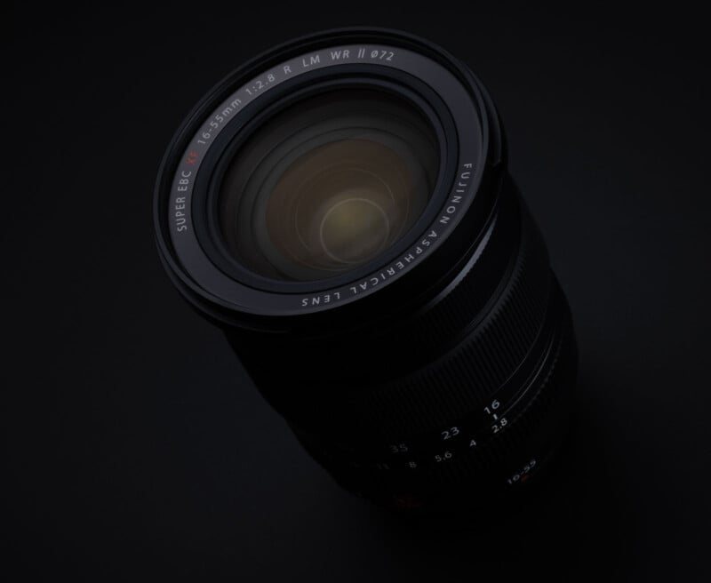 Close-up of a black Fujinon XF 16-55mm f/2.8 R LM WR camera lens placed at an angle against a dark background. The lens features visible markings for focal length and aperture settings.