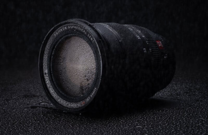 A camera lens lies on a wet surface, covered with water droplets, set against a dark backdrop.