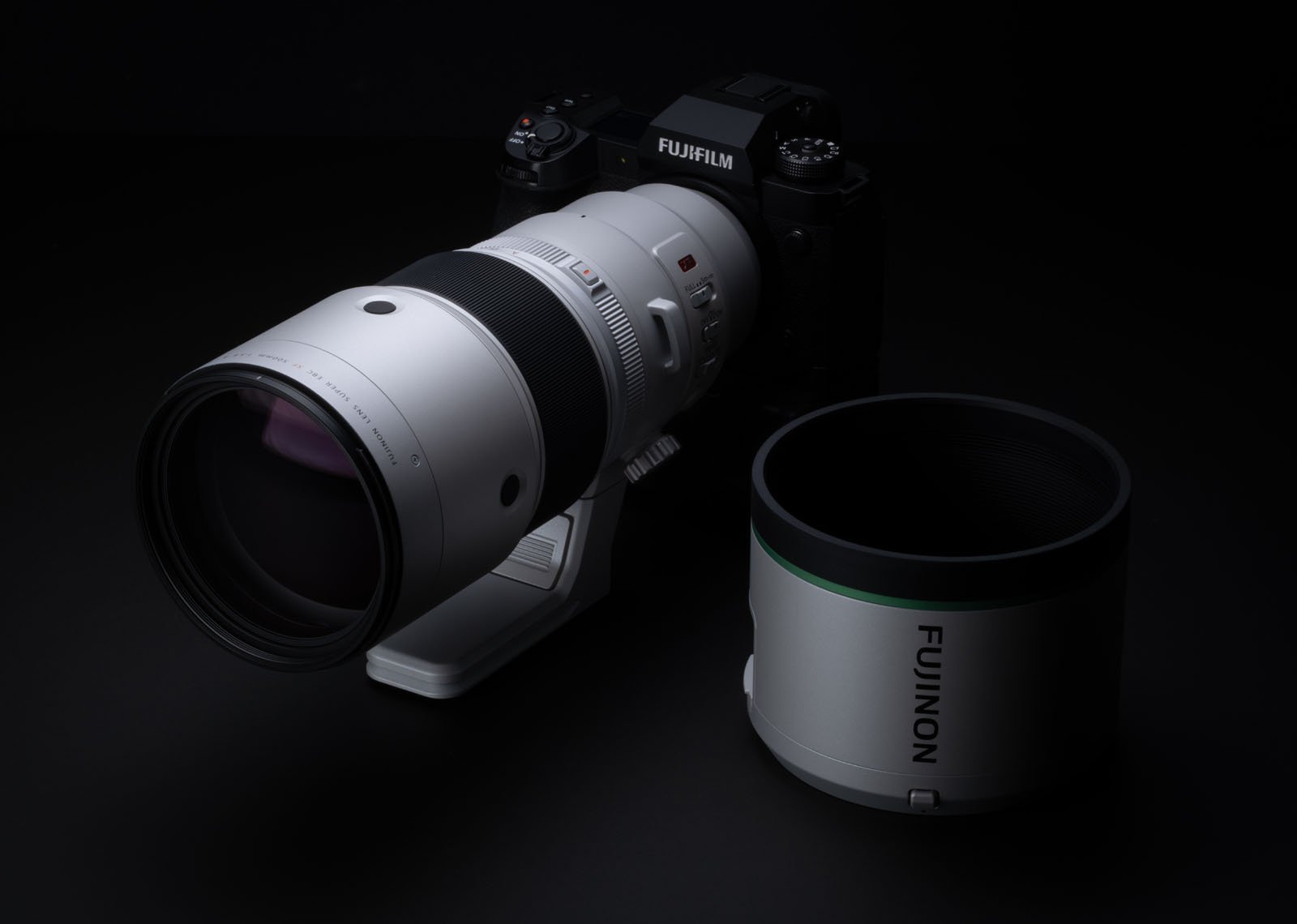 A Fujifilm camera with a large telephoto lens is displayed against a dark background. The camera is equipped with a tripod mount ring. The detachable lens hood is shown beside the camera.