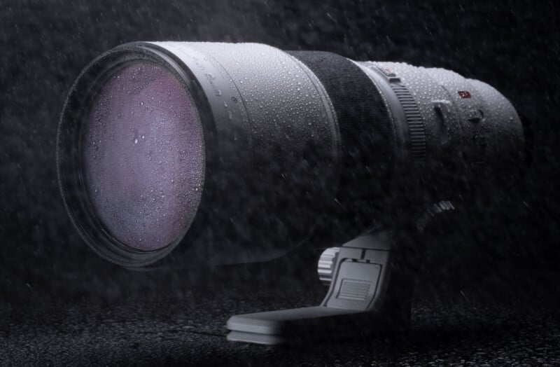 A large telephoto camera lens is positioned on a dark surface under falling rain. The lens and its body are covered with water droplets, highlighting its weather-resistant design. The backdrop is a dark, moody setting.