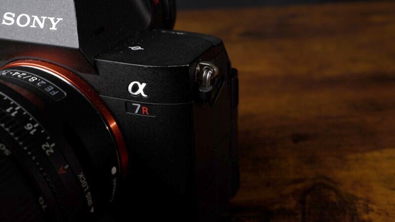 Close-up of a Sony Alpha 7R camera, showing the brand logo and red "7R" label. The camera is resting on a wooden surface, partially in shadow, highlighting its sleek, black design and lens details.