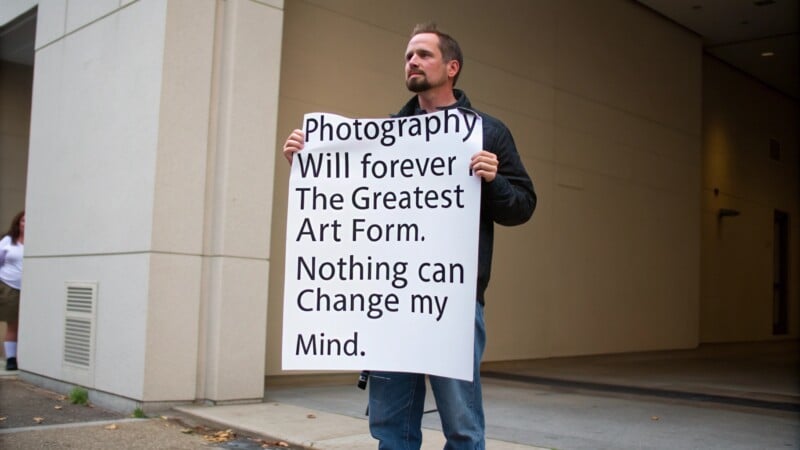 A person stands holding a large sign that reads, "Photography will forever be the greatest art form. Nothing can change my mind." They appear to be outdoors near a building.