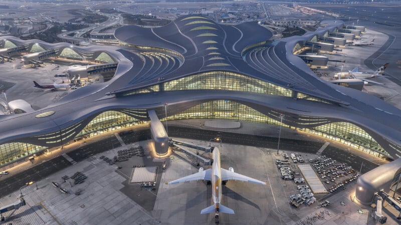 Aerial view of a modern, futuristic airport terminal with an innovative architectural design. Multiple airplanes are parked at connected gates, and the building is surrounded by roads and additional infrastructure.