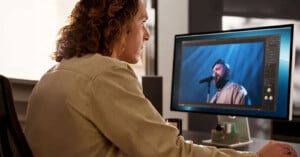 A person with curly hair sits at a desk, editing a video of a man singing into a microphone on a computer screen. The room is softly lit, with various editing tools visible on the monitor.