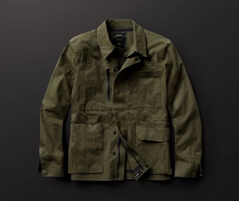 A khaki green military-style jacket with a high collar is laid flat on a dark surface. The jacket features multiple pockets and a zip front with button closures. A tag is attached to one of the pockets.