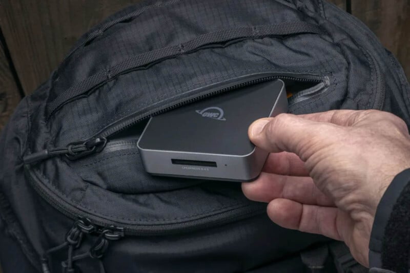 A person holds a compact, gray external hard drive with a visible logo, removing it from a black backpack. The backpack has multiple zippers and is slightly opened to reveal the tech device being retrieved.