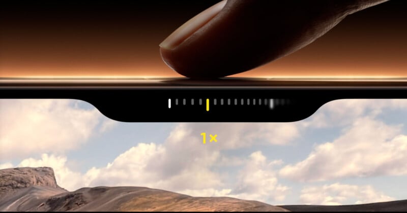 A close-up image of a finger pressing lightly on the side of a smartphone screen. The screen displays a landscape scene with mountains and cloudy sky, and shows a zoom adjustment bar at the top, indicating a 1x zoom level.