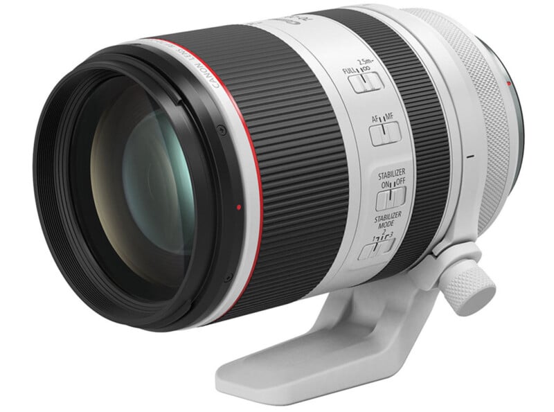 A Canon telephoto camera lens with a white and black body, featuring various switches for focus and stabilization modes. It is mounted on a white tripod collar for stability.