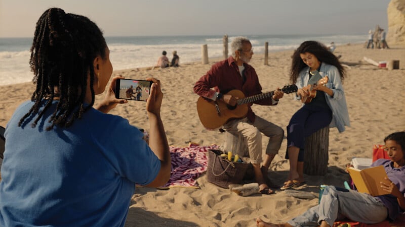 A person photographs two people playing a guitar and ukulele on a beach. They sit on logs near the ocean, with a picnic basket and a purple blanket beside them. Another person lounges on the sand, reading a book.