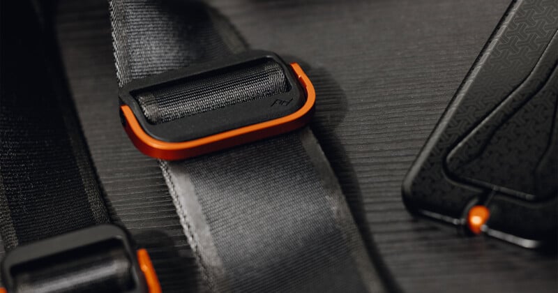 Close-up of a rugged, industrial-style strap with an orange-edged buckle on a textured dark background. The strap is secured with stitching, and a subtle pattern is visible on the surrounding material.