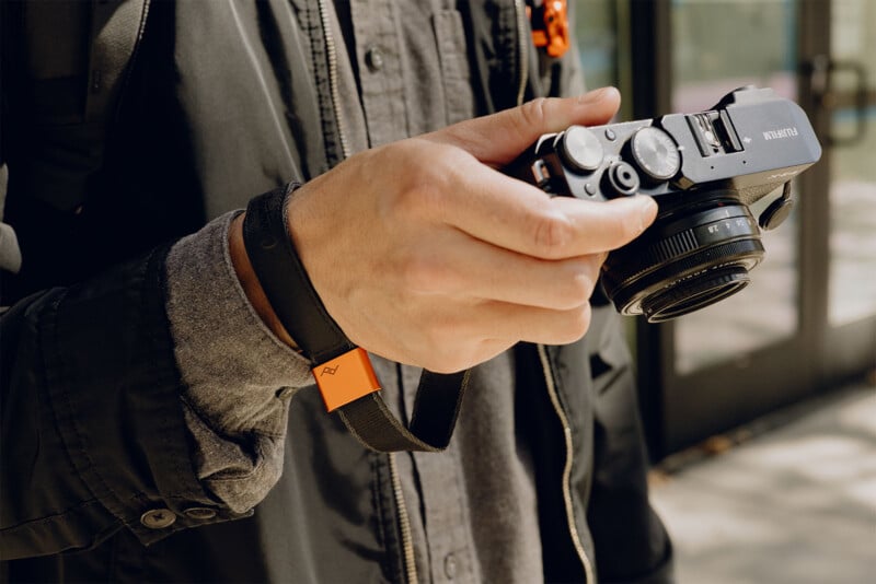 A person in a dark jacket holds a black camera with an orange and black wrist strap. The background is blurred, showing a building with glass doors. The focus is on the camera in the person's hand.