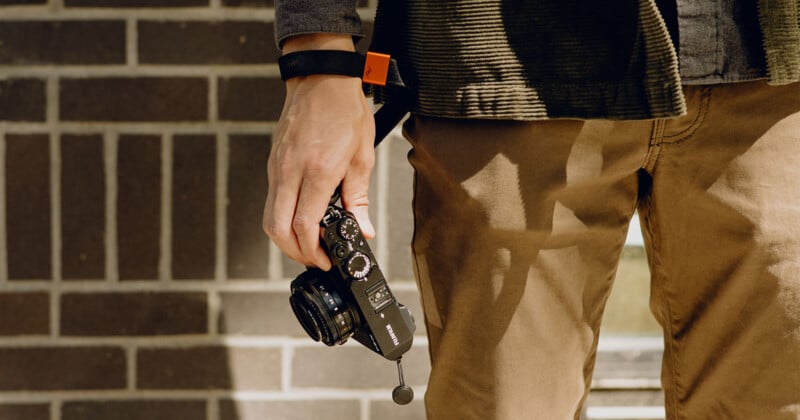 A person wearing a black watch and a dark jacket holds a black camera in their right hand. They are standing in front of a brick wall, with sunlight casting shadows on their beige pants.