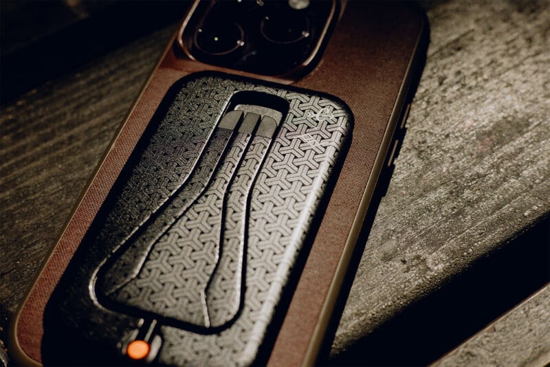 A close-up of a smartphone on a wooden surface, featuring a black leather-textured accessory with a unique pattern and an orange detail. The accessory is attached to the back of the phone, partially covering the camera lenses.