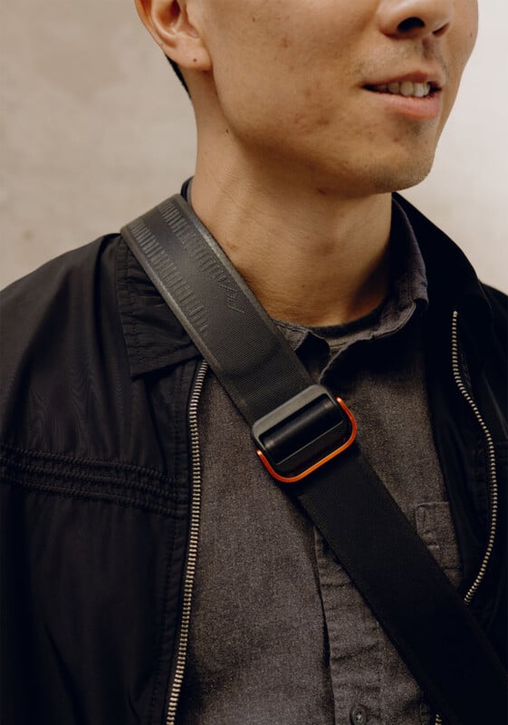 A person wearing a black jacket and a dark shirt, with a black crossbody strap over their shoulder. The strap has a metal buckle with orange accents. The image focuses on the upper half of the person.