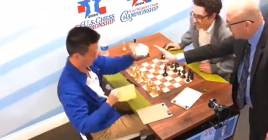 A chess player in a blue jacket raises his arm while another player watches, seated opposite him at a chessboard. An official in a suit points at the board. The backdrop displays "U.S. Chess Championship 2023" logos.