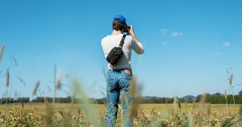 A person wearing a blue cap and jeans stands in a field taking photos with a camera against a clear blue sky. They have a black crossbody bag and the background shows a vast expanse of grass and distant trees.