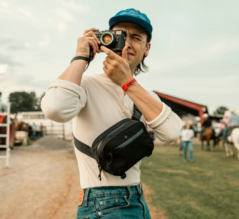 A person wearing a blue cap and white shirt is taking a photo with an analog camera. They have a black crossbody bag and are standing outdoors, with horses and people in the background. The scene has a casual, relaxed atmosphere.