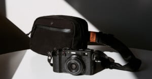 A black, vintage-style camera with a strap is placed on a white surface. Next to it is a black rectangular camera bag. Sunlight and shadows create a contrasting background.