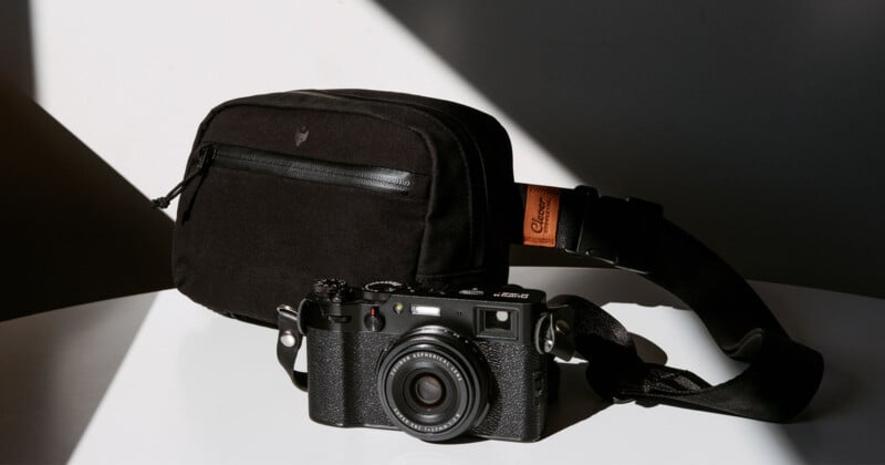 A black, vintage-style camera with a strap is placed on a white surface. Next to it is a black rectangular camera bag. Sunlight and shadows create a contrasting background.