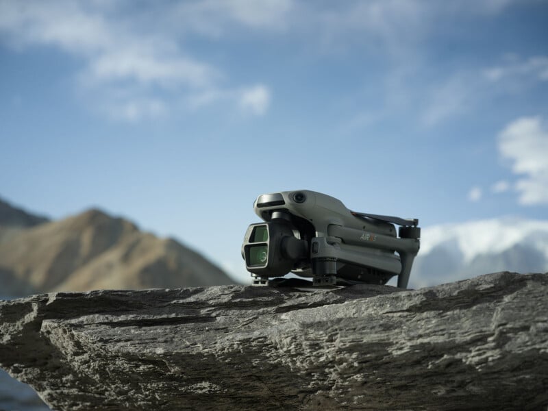 A drone sits on a large rock against a backdrop of mountains and a partly cloudy sky, with sunlight illuminating the scene.