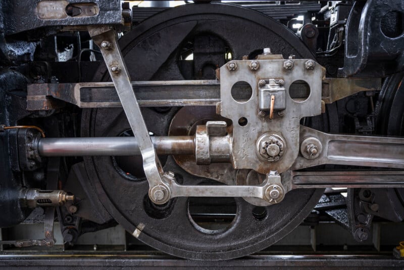 Close-up image of the intricate inner workings of a steam locomotive's driving wheel and connecting rods. The metallic components, gears, and bolts are prominently displayed, showcasing the complex engineering and mechanical details.