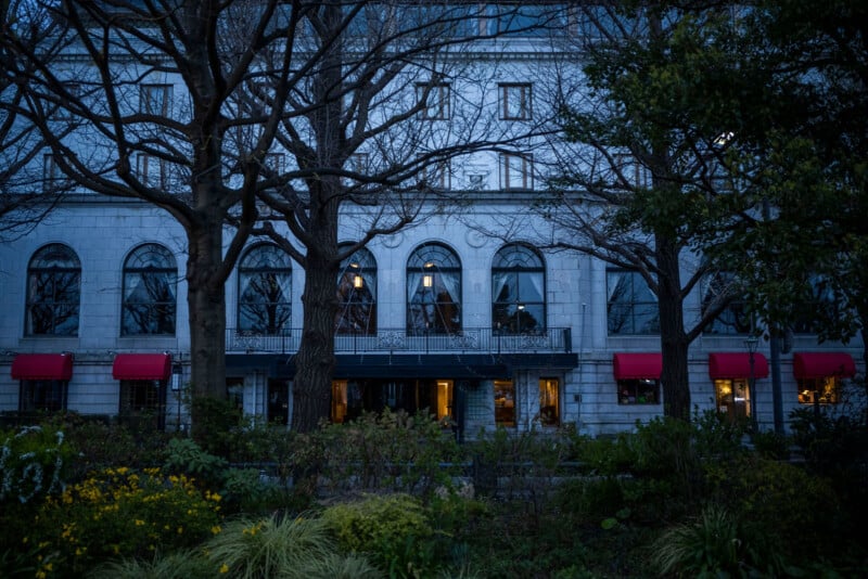 A stately, gray stone building with five large, arched windows and red awnings is fronted by a garden with bare trees and greenery in the foreground. The scene is dimly lit, suggesting evening or early night.