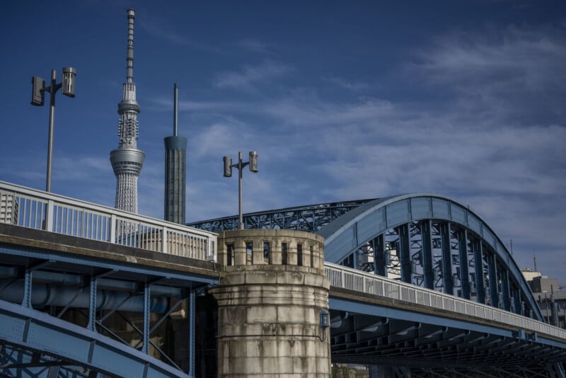 A blue steel bridge with a rounded arch spans across the image. In the background, the Tokyo Skytree tower rises against a partly cloudy sky. The bridge features streetlights and structural supports, with a stone pillar at one end for added architectural detail.