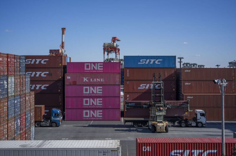 Image showing a bustling shipping container yard under a clear blue sky. Stacks of large shipping containers from various companies, prominently featuring the word "ONE," are arranged. Trucks and a container handler are actively organizing the containers.