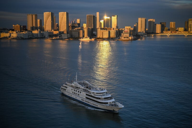 A large white yacht sails across a calm harbor at sunset with a modern city skyline illuminated by the setting sun in the background. The golden light creates a reflection on the water, enhancing the serene atmosphere.