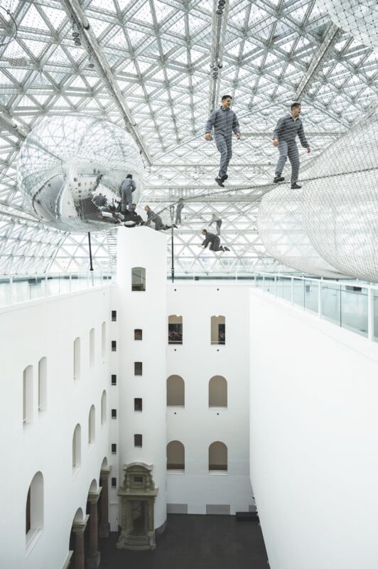 People walking on transparent, netted walkways suspended inside a modern building with a geometric glass ceiling. The interior features white walls and arched windows, creating a bright and airy atmosphere.