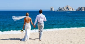 A bride and groom are walking hand in hand on a sandy beach. The bride wears a white dress with a veil, holding a bouquet. The groom is in a white shirt and gray pants. In the background is a vast blue ocean with distant rocks.