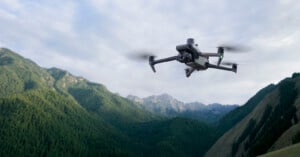 A drone flies against a backdrop of lush, green mountains under a cloudy sky. Its rotors are in motion as it hovers in the air, capturing the rugged landscape.