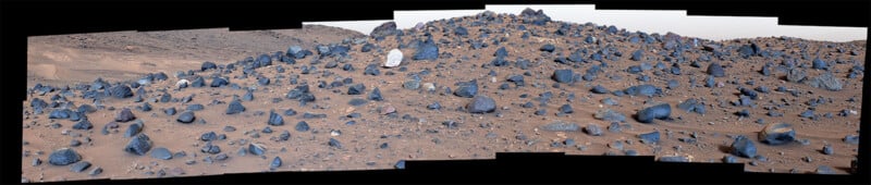 Panoramic image of a rocky Martian landscape featuring numerous scattered dark rocks and pebbles across reddish-brown sand. The terrain is uneven, with small hills, under a pale sky. No vegetation or water is visible.