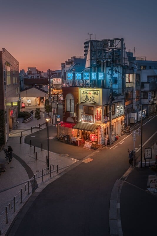 A twilight view of a bustling urban corner in Japan, featuring a multi-story building adorned with colorful, illuminated signs. The street is lined with shops and restaurants, creating a cozy, vibrant atmosphere against a dusky sky.