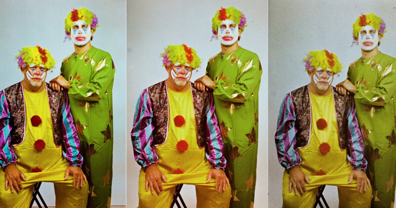 Two people are dressed as clowns in this image. The seated clown wears a yellow outfit with a multicolored vest and red buttons, while the standing clown wears a green suit with star patterns. Both have colorful wigs and face paint.