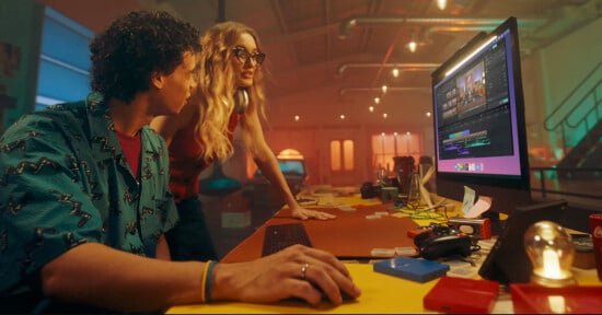 Two people collaborate at a desk in a vibrant office. One person, seated, operates a computer with editing software on the screen. The other, standing, leans over while discussing the project. The room is warmly lit, featuring colorful decor.