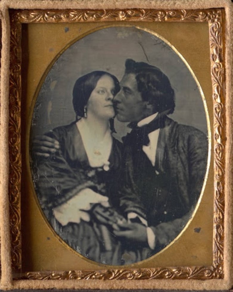 A vintage photograph in an ornate frame shows a man and woman in 19th-century attire, seated closely. The man leans in with one arm around the woman, who gazes at him, creating an intimate and affectionate scene.