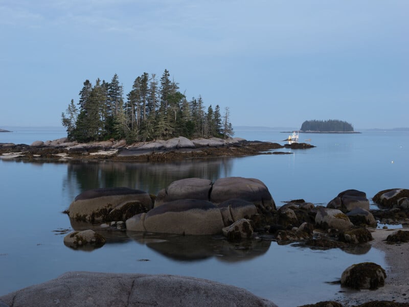 A serene coastal scene featuring a small rocky island with tall evergreen trees, surrounded by calm water. In the distance, another island is visible. The sky is overcast, casting a tranquil atmosphere over the landscape.