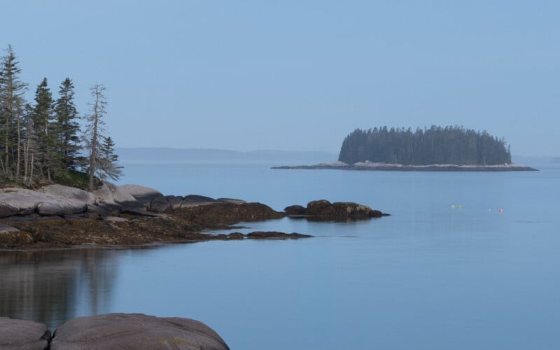 A serene coastal scene featuring calm blue waters, rocky shores with sparse trees on the left, and a small, tree-covered island in the distance under a pale blue sky. Three buoys float on the water.